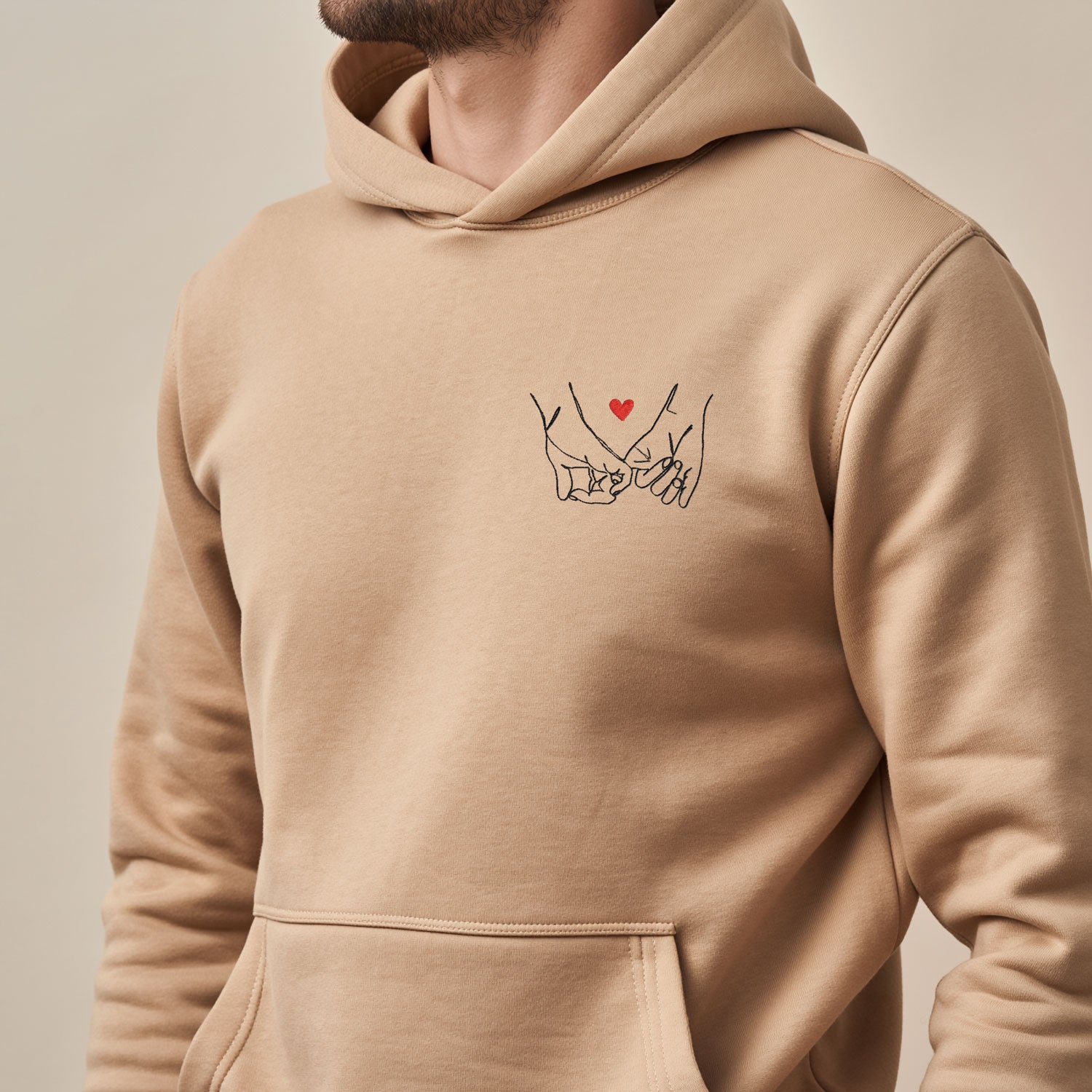 Sweat à capuche sable clair brodé avec motif mains entrelacées et petit cœur rouge, porté par un adulte, broderie positionnée côté cœur, style couple homme femme