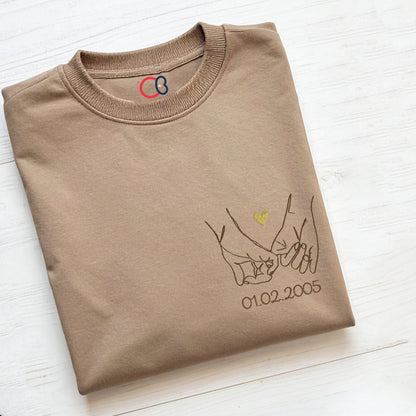 T-shirt beige brodé avec motif de mains entrelacées en line art, petit cœur doré et date personnalisée, idée cadeau Saint-Valentin ou anniversaire de couple.