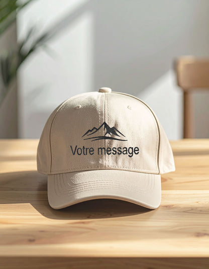 Casquette beige personnalisée en coton brossé, posée de face sur une table en bois clair avec broderie message et motif montagne.